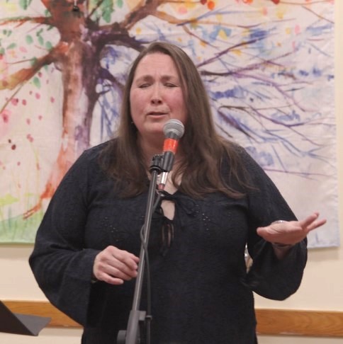 An image of Esther Rhatigan singing in front of a painting of a tree with rainbow colored leaves - artist unknown. Photo credit: Kevin Kwasny